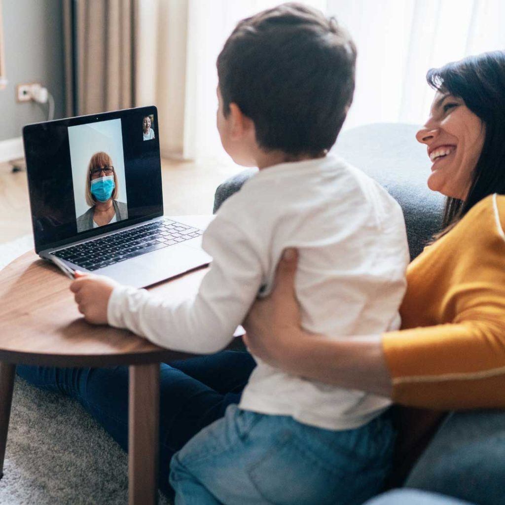 mother and boy sitting in front of laptop video chatting with woman wearing mask