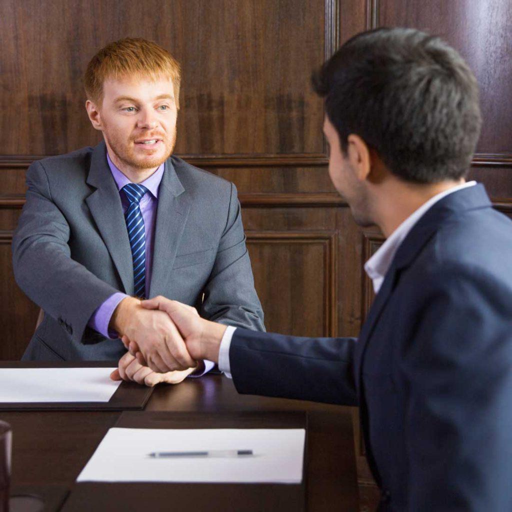 man in red hair wearing grey suit sitting at table shaking hands with man in dark suit