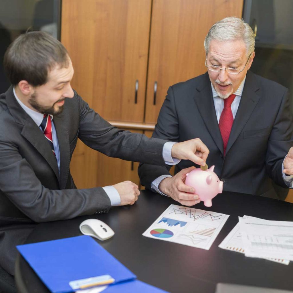 younger man in suit placing coin into piggy bank held by older man in suit