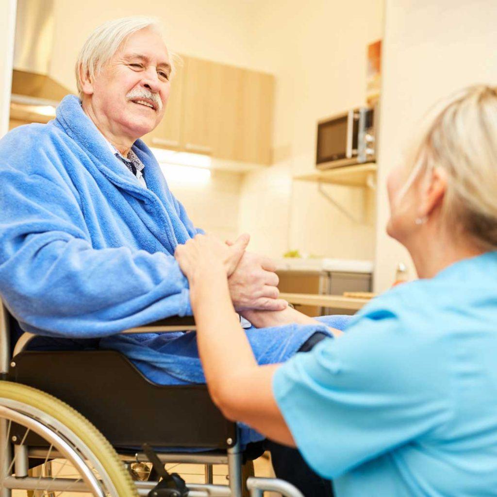 older man in blue robe sitting in wheel chair with woman in blue shirt placing her hand over his