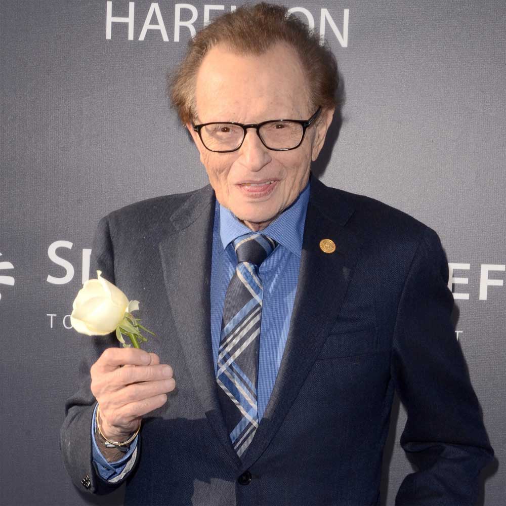 larry king in a black suit with a blue shirt on holding a white rose