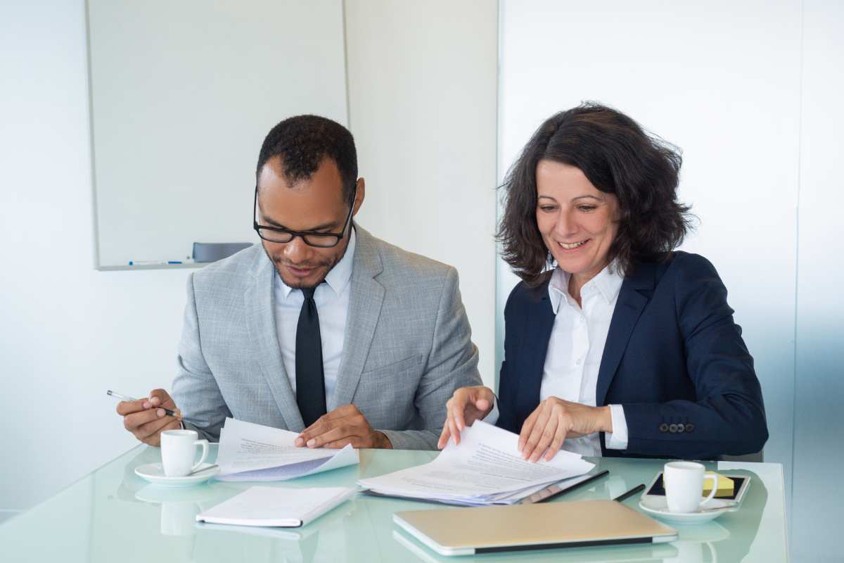 man and woman in suits sitting at the end of a table going through documents