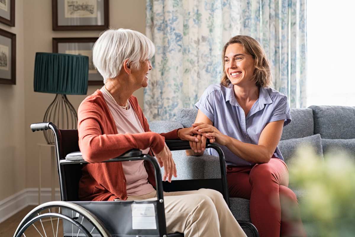 smiling woman sitting on a couch holding the hand of an elderly woman in a wheelchair
